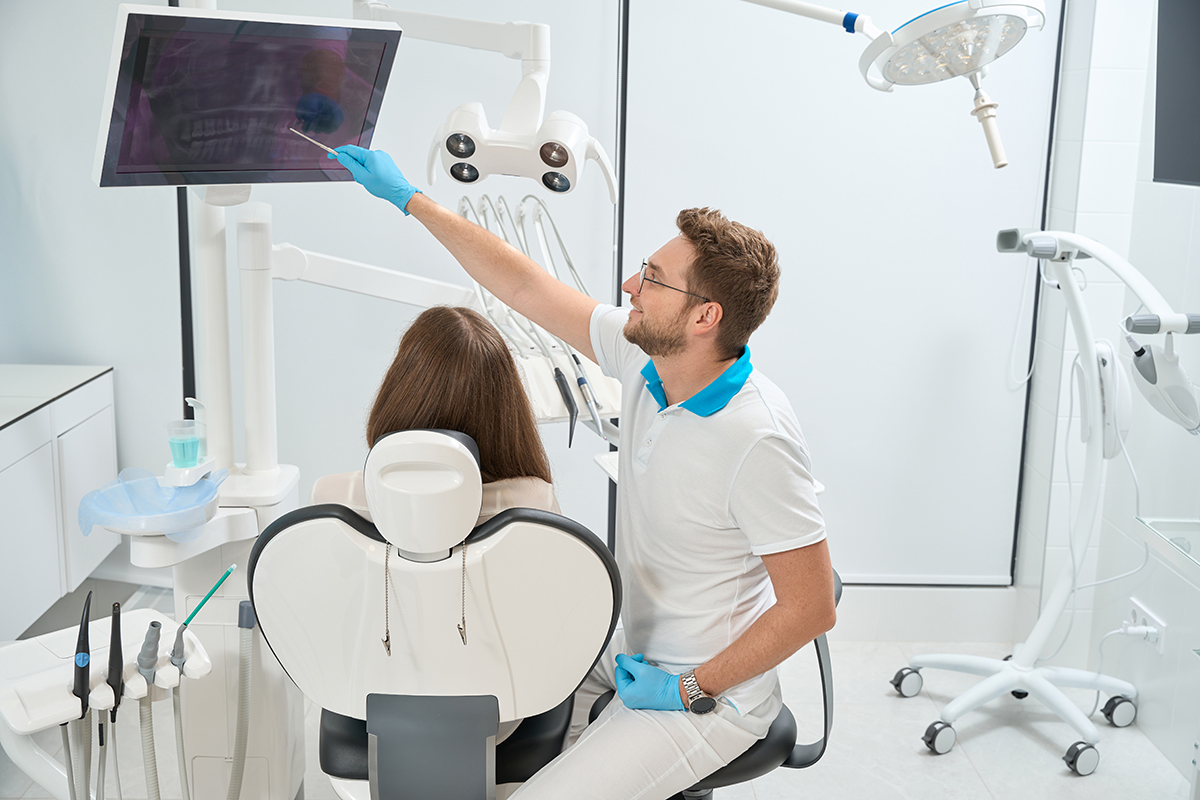 Dentist pointing at problem tooth on digital panoramic x-ray on computer screen to female client