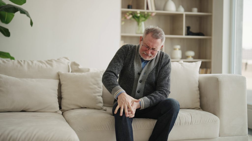 Senior man sitting on a couch at home holding his knee in pain, suffering from joint problems.